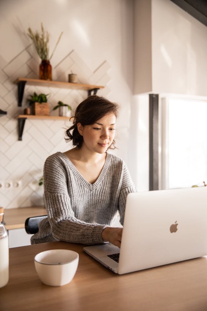 hero-gallery-03 Young female in gray sweater sitting at wooden desk with laptop and bottle of milk near white bowl while browsing internet on laptop during free time at home