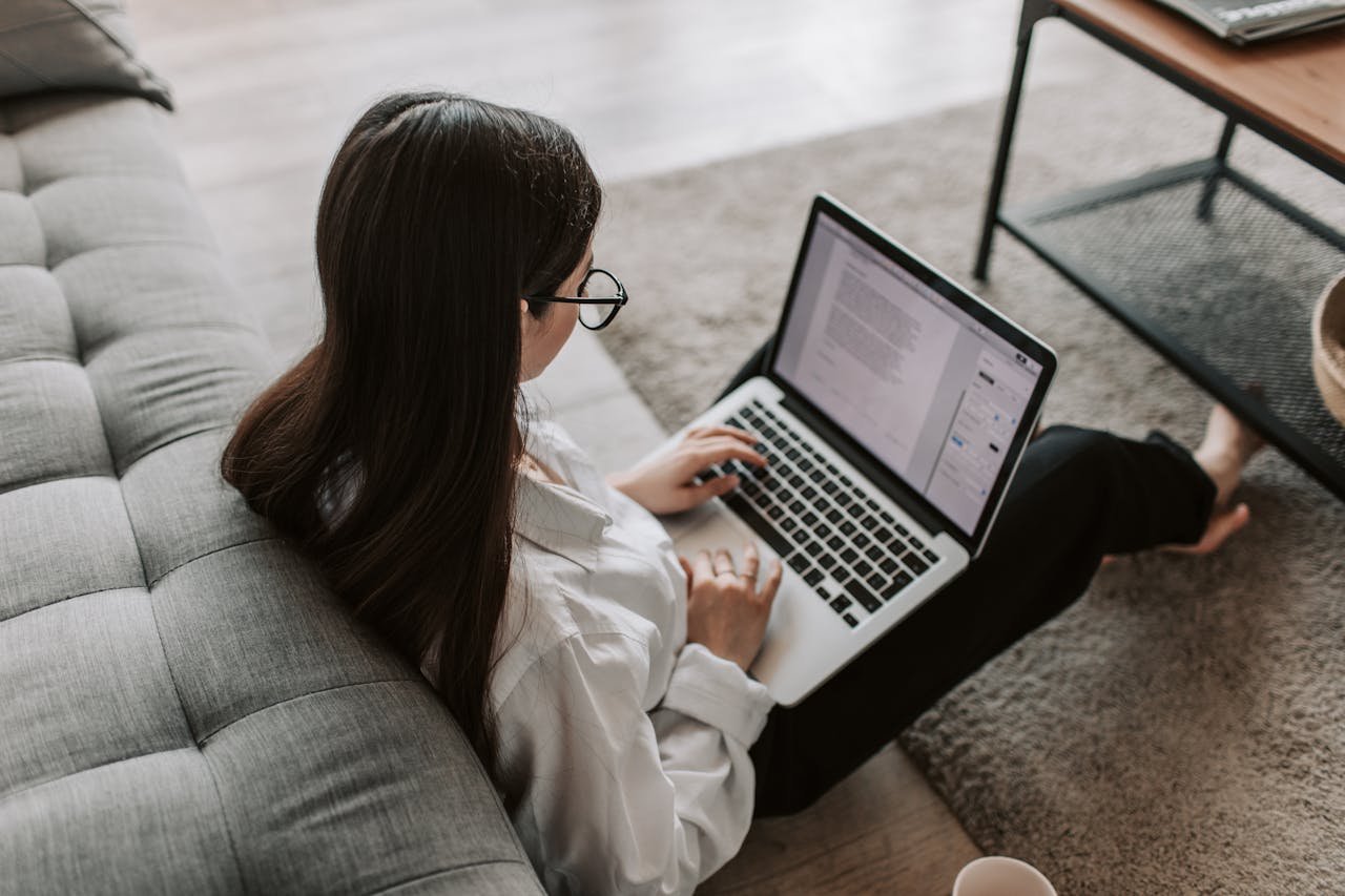 hero-gallery-01 Woman working remotely with a laptop on the floor next to a sofa, enjoying comfortable home office setup.